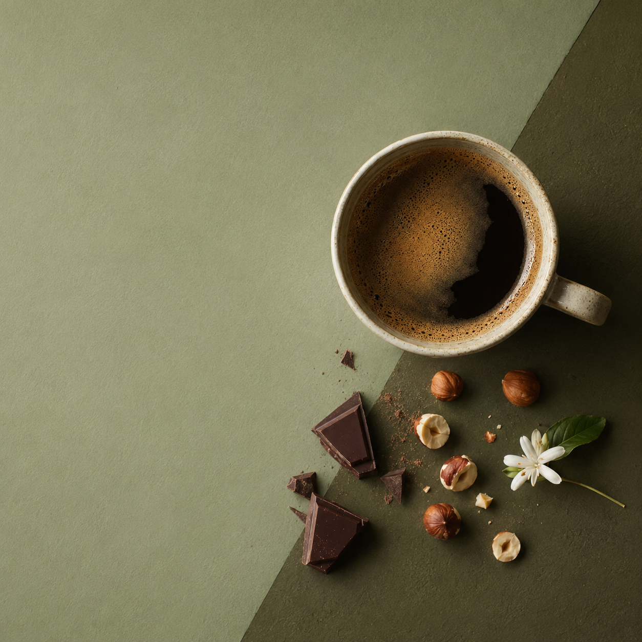 A cup of coffee on an olive green surface with dark chocolate pieces, hazelnuts and a white coffee blossom